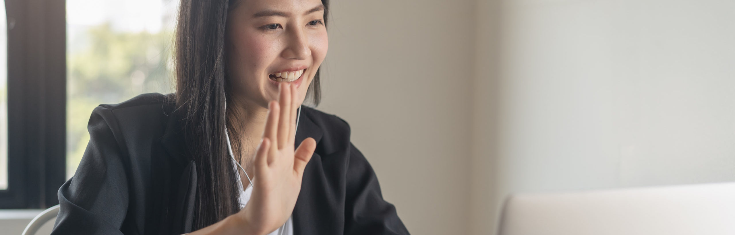 Happy young Asian business woman waving hands to greeting partner during making video conference with her team.