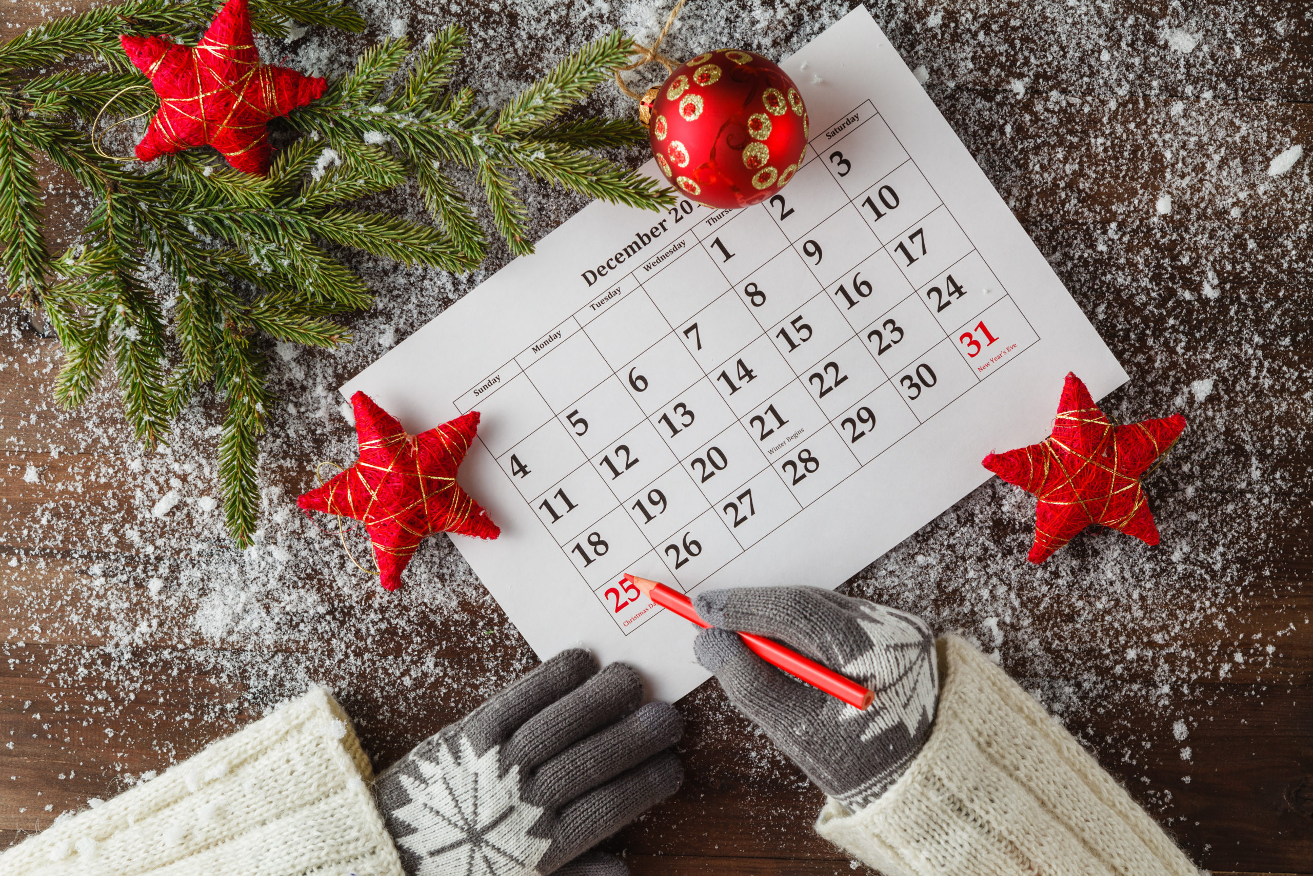 Person wearing snowflake gloves writing on a December calendar surrounded by holiday decorations